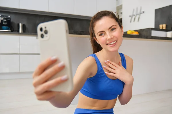 Smiling beautiful girl records video while doing sports at home, looks at smartphone, takes selfie on mobile phone, workout indoors in blue leggings and sportsbra.