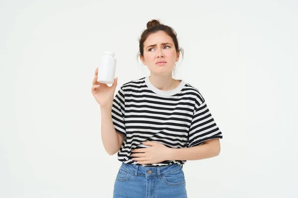 Image of woman with stomach ache, shows bottle with pills, takes tablet from menstrual cramps, period pain, soothing discomfort, standing over white background.