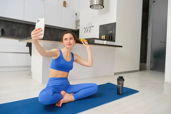 Portrait of active girl, vlogger does sports, records her workout training from home on smartphone camera, posing for selfie inside her house, sits on rubber yoga mat in blue leggings and sportsbra.
