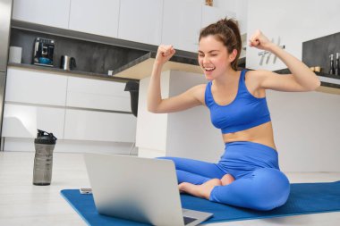 Heyecanlı genç bir kadının portresi, dizüstü bilgisayarla minderde oturmak, müşteriye tezahürat yapmak, online yoga dersi vermek, uzaktan fitness dersi vermek, evden beden eğitimi öğretmeni olarak çalışmak..