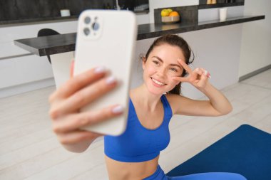Kızın portresi, spor yaparken akıllı telefondan selfie çekiyor. Genç kadın egzersizlerini evde yapıyor, spor blogu için selfie çekiyor, oturma odasında oturuyor..
