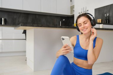 Genç mutlu kadının portresi kulaklıkla egzersiz yapıyor, yoga yaparken müzik dinliyor, mavi spor kıyafetleriyle lastik mindere oturuyor..