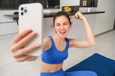 Genç enerjik kız evde spor yapıyor, kolundaki kaslarını gösteriyor ve akıllı telefondan selfie çekiyor oturma odasındaki spor paspasında poz veriyor..