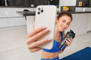 Sporcu kadının portresi oturma odasında su şişesiyle selfie çeker, akıllı telefon tutar ve spor yaparken poz verir..