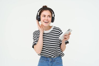 Akıllı telefonlu mutlu bir kadının portresi cep telefonuyla şarkı değiştiriyor, kulaklıkla müzik dinliyor, beyaz arka planda dans ediyor..