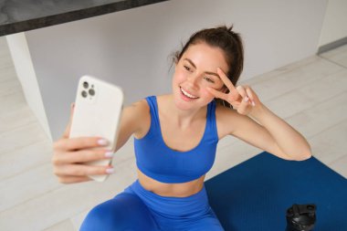 Güzel bir bayan sporcunun portresi, evde yoga minderiyle egzersiz yapan bir kadın, akıllı telefondan selfie çekiyor, spor eğitmeninin sosyal medyası için fotoğraflar çekiyor..