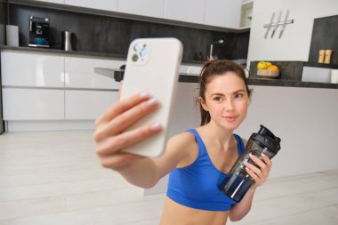 Genç fitness kızının portresi, akıllı telefondan su şişesiyle selfie çekiyor, antreman sırasında su içiyor, evde eğitim görüyor, spor videolarını kaydediyor..