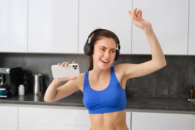 Güzel bir spor kadının portresi, aktif giysi giymesi, mutfakta kulaklıkla dans etmesi, müzik dinlemesi ve eğlenmesi, antrenmandan sonra enerji hissetmesi..