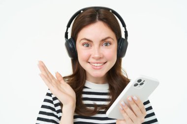 Güzel bir genç kadının portresi, kulaklık takıyor, akıllı telefon tutuyor, gülümsüyor ve mutlu görünüyor, beyaz arka planda izole edilmiş..