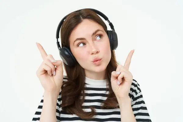 Portrait of young brunette woman in headphones, listening music in headphones, pointing fingers up, showing banner, advertisement, isolated over white background.
