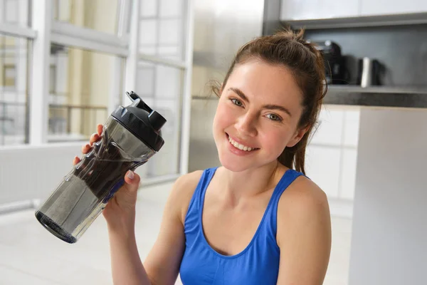 Smiling fitness girl drinks water in the middle of training session, smiling and looking happy, workout from home in blue sportsbra.