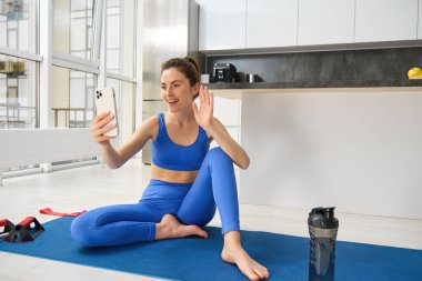 Güzel, atletik bir kız görüntüsü evde yoga minderinde oturuyor, akıllı telefonuyla selfie çekiyor ve egzersiz yapıyor, spor yapıyor..