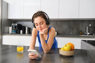 Mutfakta dikilip portakal suyu içen, kulaklıkla müzik dinleyen, akıllı telefon uygulaması kullanan mutlu, şık bir spor kadını görüntüsü..