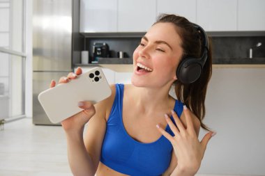Mutlu, güzel bir fitness kadınının portresi, evde egzersiz yapmak, kablosuz kulaklık takmak ve akıllı telefon başında şarkı söylemek..