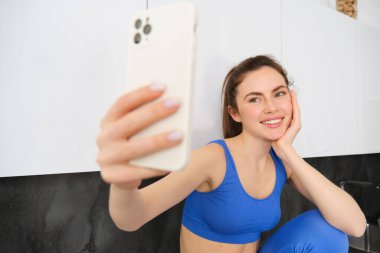 Fotograf için fitness kızı portresi, akıllı telefon uygulamasında selfie çekmek, mutfakta oturmak, aktif giysi giymek..
