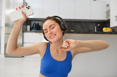 Mutluluk, spor ve refah. Genç bir kadın kulaklıkla dans ediyor, elinde akıllı telefon, evinde egzersiz yapıyor, evinde spor yapıyor..