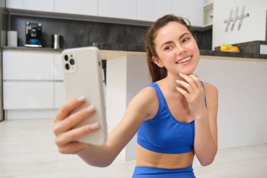 Genç fitness kızı portresi, blogcu evden antrenmanını kaydediyor, akıllı telefonda selfie için poz veriyor, kamerada yoga yapıyor, mavi spor sutyen ve taytla oturuyor..
