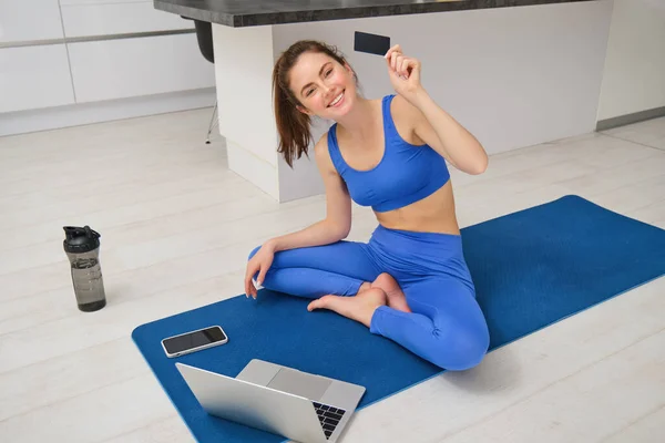 Portrait of young fitness girl paying for online class subscription, workout app, holding credit card, sitting on yoga mat with laptop, buying fitness tutorials.