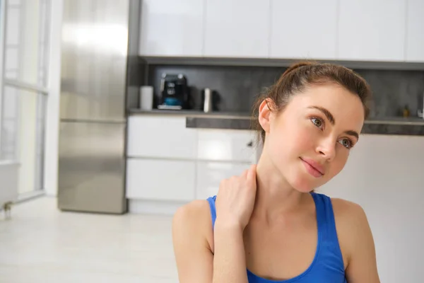 Close up portrait of young woman does fitness at home, touches her neck, rubs shoulders after workout training session. Wellbeing and sport concept