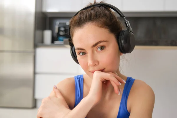 Close up portrait of brunette fitness girl, listens music in wireless headphones, smiles at camera, does workout alone at home, sits on floor and exercises.