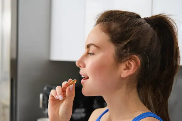 Close up portrait of fitness girl, taking vitamins, holding pill dietary supplement, swallowing tablet buds for healthy and strong body, standing in kitchen.