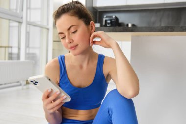 Akıllı telefonlu genç bir kadın evde spor yapmak için cep telefonu uygulaması kullanıyor. Spor eğitimi için evde, mavi spor sutyen ve tayt giyiyor..