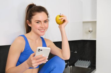 Fitness kadınının portresi, akıllı telefonuyla mutfakta oturmak, sosyal medyayı kaydırmak ve elma yemek, egzersiz arasında sağlıklı bir şeyler yemek, spor kıyafetleri giymek..