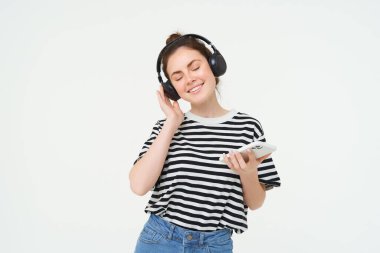 Akıllı telefonlu genç bir kadın müzik dinliyor, kulaklıkla en sevdiği şarkıyla dans ediyor, beyaz arka planda poz veriyor..