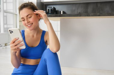 Mavi spor ayakkabılı ve tozlu güzel bir kadın resmi. Akıllı telefonuna bakıyor, egzersiz yapıyor, antrenmanlarını kaydediyor..
