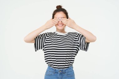 Sürprizi bekleyen bir kızın portresi, ellerle gözleri kaplayan, parmaklarından bakan, beyaz arka planda duran.