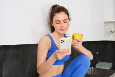 Fitness kadınının portresi, akıllı telefonuyla mutfakta oturmak, sosyal medyayı kaydırmak ve elma yemek, egzersiz arasında sağlıklı bir şeyler yemek, spor kıyafetleri giymek..