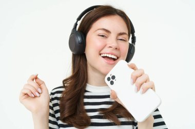 Teknoloji ve sosyal medya. Karaoke yapan mutlu genç bir kadının portresi, akıllı telefonda müzik uygulaması çalan, kablosuz kulaklık takan, beyaz arka planda duran.