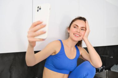 Formda ve sağlıklı, gülümseyen fitness kızı tasviri, cep telefonuyla selfie 'ye poz vermek, akıllı telefon tutmak, ekrana bakmak, spor ve taytla fotoğraf çekmek..