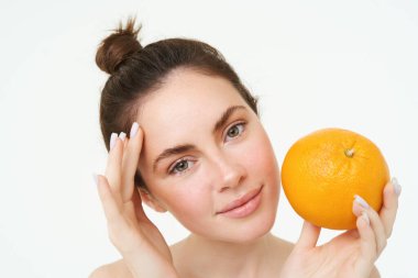 Esmer kadın modelin yakın portresi, turuncuyu yüzüne yakın tutuyor, beyaz arka planda duruyor..