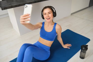 Kablosuz kulaklıklı kadın portresi, fitness minderinde oturur, antrenman sırasında selfie çeker ve fotoğraf çeker, evde egzersiz yapar..