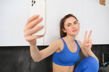 Formda ve sağlıklı genç spor kadını, barış işareti gösteriyor, aktif giyimde selfie çekiyor, spor blogcusu sosyal medya için aktivite giysilerinde fotoğraflar çekiyor..