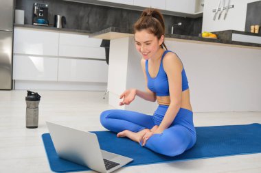 Genç fitness eğitmeninin çevrimiçi antrenman için dizüstü bilgisayar kullanması, spor egzersizleri göstermesi, müşteriyle konuşması, spor kıyafetlerinde yoga kauçuk minderinde oturması..