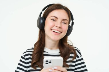 Güzel genç bir kadının portresi kablosuz kulaklıkla müzik dinliyor, cep telefonunu kullanıyor, gülümsüyor ve mutlu görünüyor, beyaz arka planda izole edilmiş..