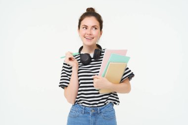 Boynunda not defterleri ve kulaklıkları olan, üniversite reklamı için poz veren beyaz arka planı olan genç bir kadının portresi..