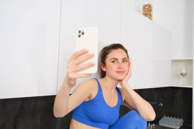 Fotograf için fitness kızı portresi, akıllı telefon uygulamasında selfie çekmek, mutfakta oturmak, aktif giysi giymek..