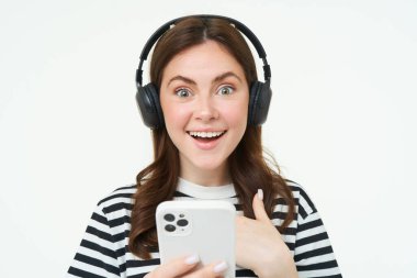 Şaşırmış yüzlü genç bir kadın, akıllı telefon ekranına bakıyor, şaşırıyor, cep telefonunda mutlulukla bakıyor, kablosuz kulaklıklar takıyor, beyaz arka plan.