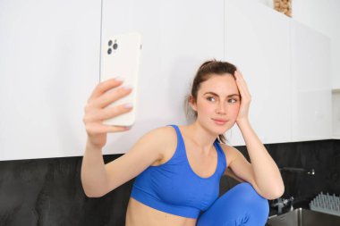 Güzel bir sporcunun portresi, spor sutyen ve tayt giyiyor, mutfakta oturuyor ve fotoğraf çekiyor, akıllı telefon uygulamasında poz veriyor..