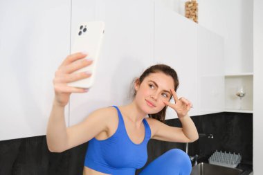 Gülümseme portresi, spor salonu eğitmeni fitness blogu için poz verme, akıllı telefonda selfie çekme, mutfakta oturma, mutlu görünme..