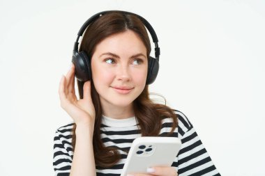 Mutlu genç kadın portresi, kablosuz kulaklıklı kadın model, gülümseyen ve kameraya bakan, akıllı telefon tutan, cep telefonu uygulaması kullanan, beyaz arka plan.