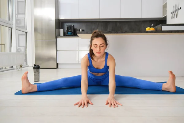 Portrait of young woman does gymnastics training at home, workout, making splits on rubber fitness mat, sitting on floor with feet stretched sideways.