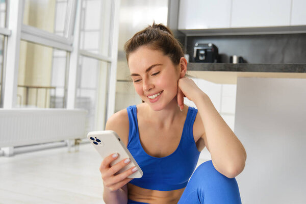 Image of young fit woman wearing sportbra and blue leggings, using smartphone app, does workout from home, sits on floor and follows online training video.