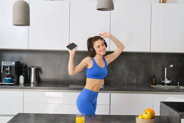 Portrait of beautiful fitness blogger, woman in headphones, listening music and dancing in kitchen, wearing blue leggings and sportsbra.