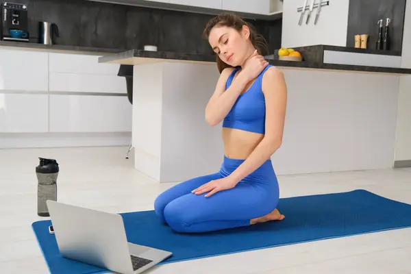 Image of calm and relaxed woman, sitting on yoga mat with laptop, massaging her neck, warm-up body before workout training, home fitness session.