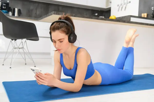 Image of young woman watches sport gym tutorials on smartphone, laying on yoga mat, doing fitness workout at home, wearing blue leggings and sportsbra.