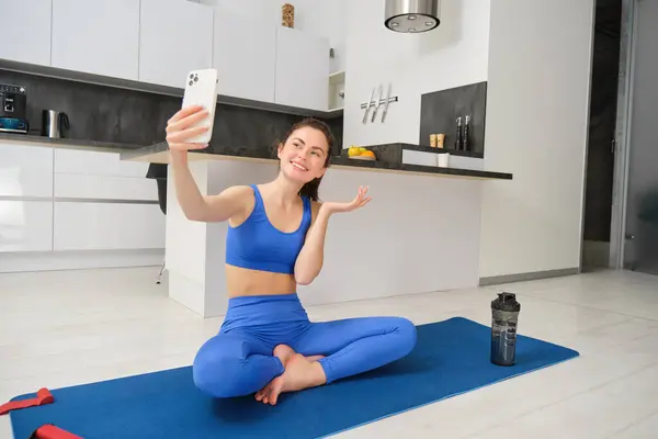 Active young woman does sports, records her workout training from home on smartphone camera, posing for selfie inside her house, sits on rubber yoga mat in blue leggings and sportsbra.